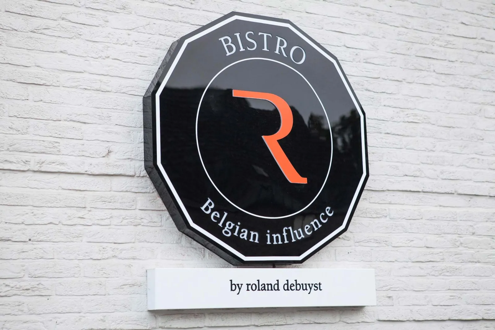 Het bord van Bistro R met de tekst “belgian influence, by roland debuyst” op een witte muur